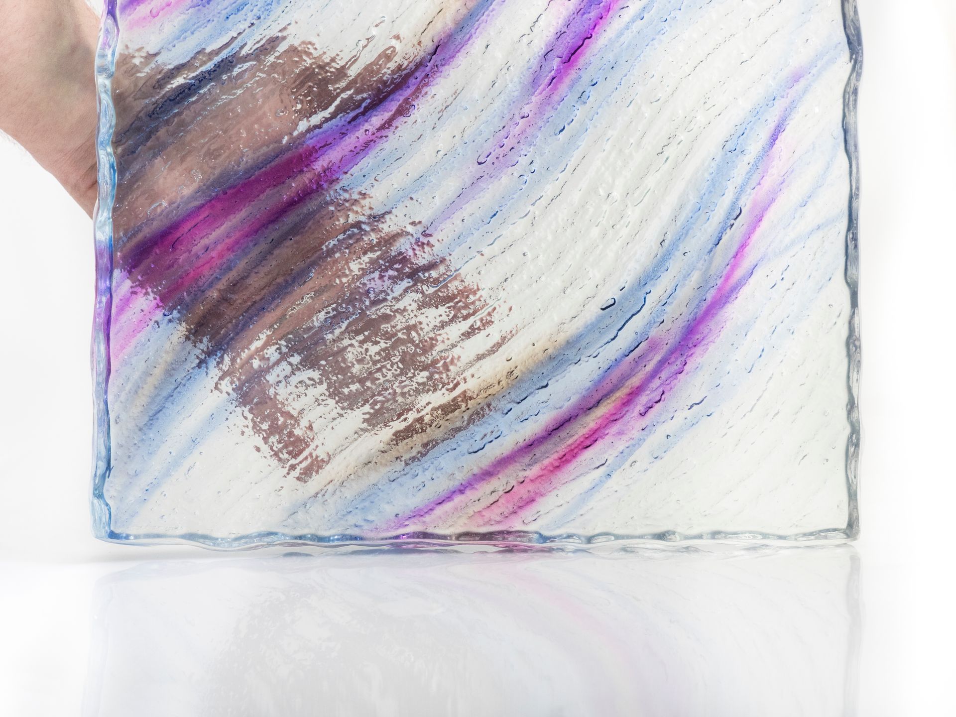 A hand holds a square, textured surface featuring abstract, diagonal streaks of purple, blue, and brown on a white base.