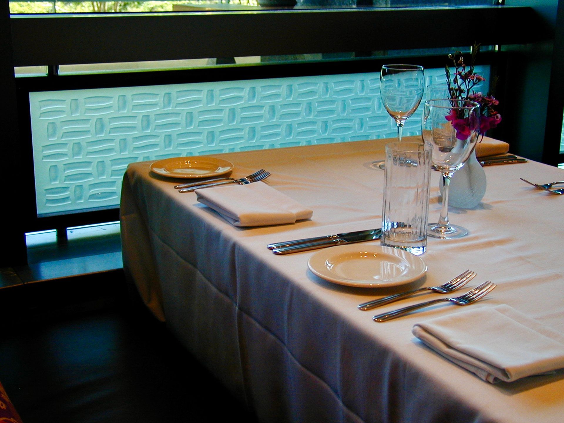 A formal restaurant table setting with white linens, napkins, silverware, glassware, and a small vase of flowers.