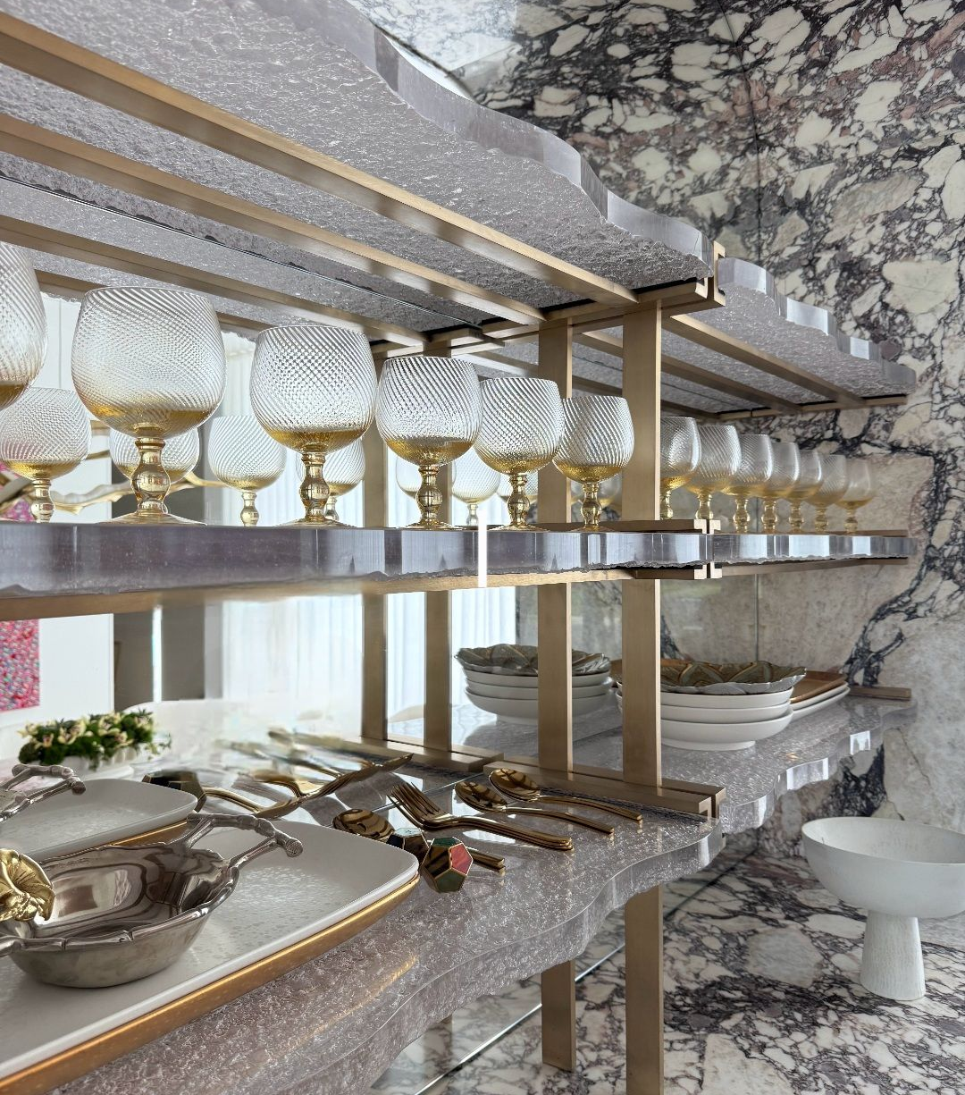 Elegant tableware on cast glass shelves against a marbled wall; includes glasses, silverware, and serving dishes.