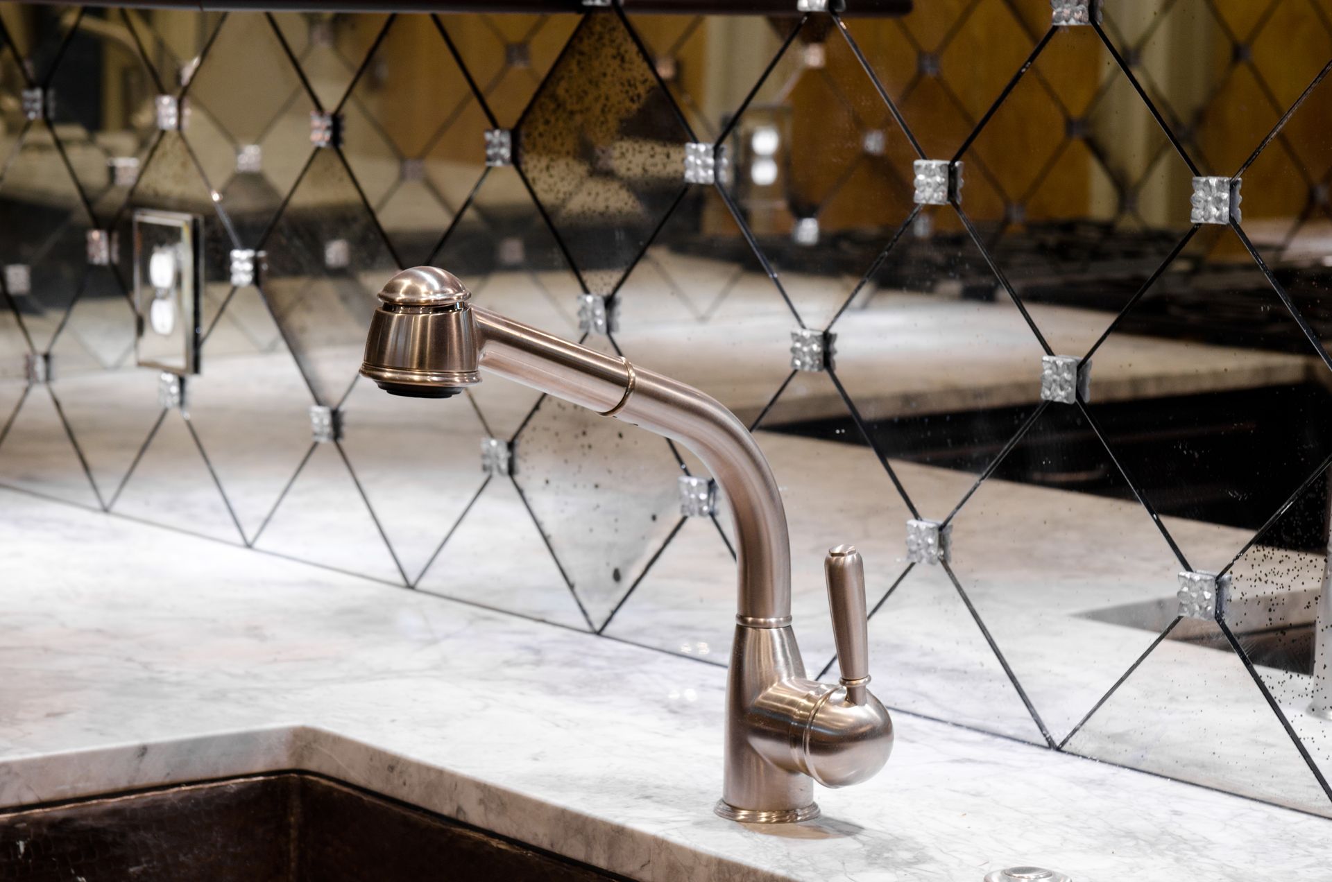 A stainless steel kitchen faucet with a pull-down sprayer sits on a marble countertop in front of a mirrored backsplash.