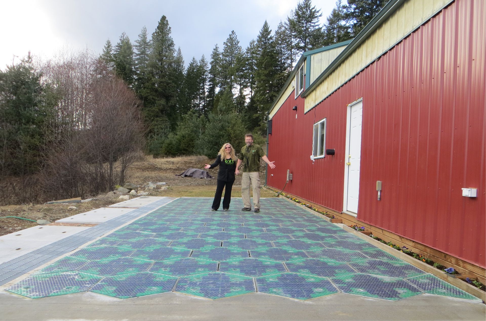 Two people stand on a large, patterned blue and green mosaic glass floor surface next to a red barn.