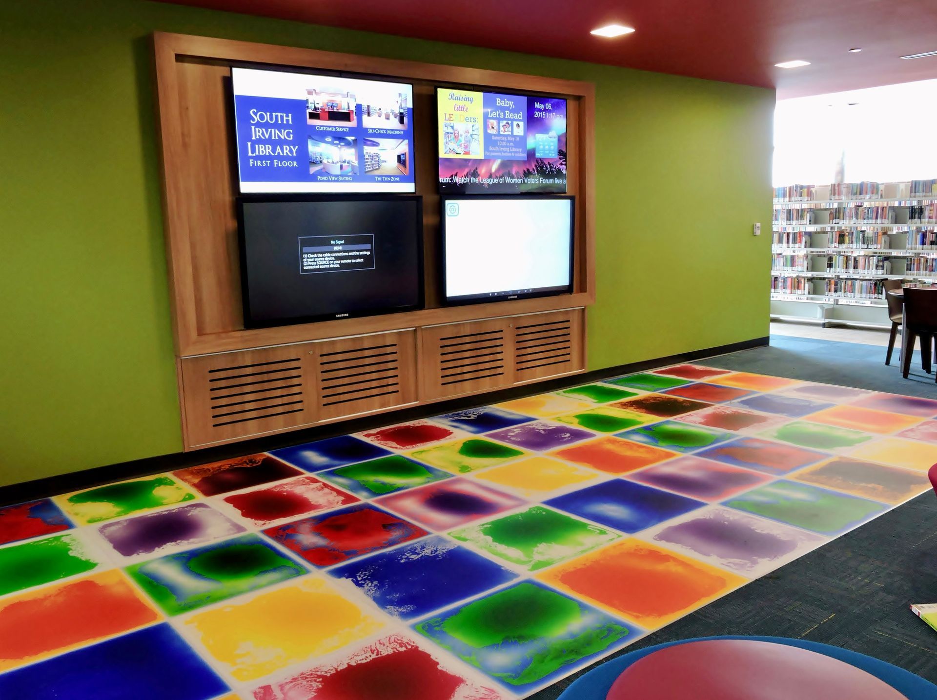 Four television screens mounted on a wooden wall above a floor with colorful, translucent square tiles in a library.