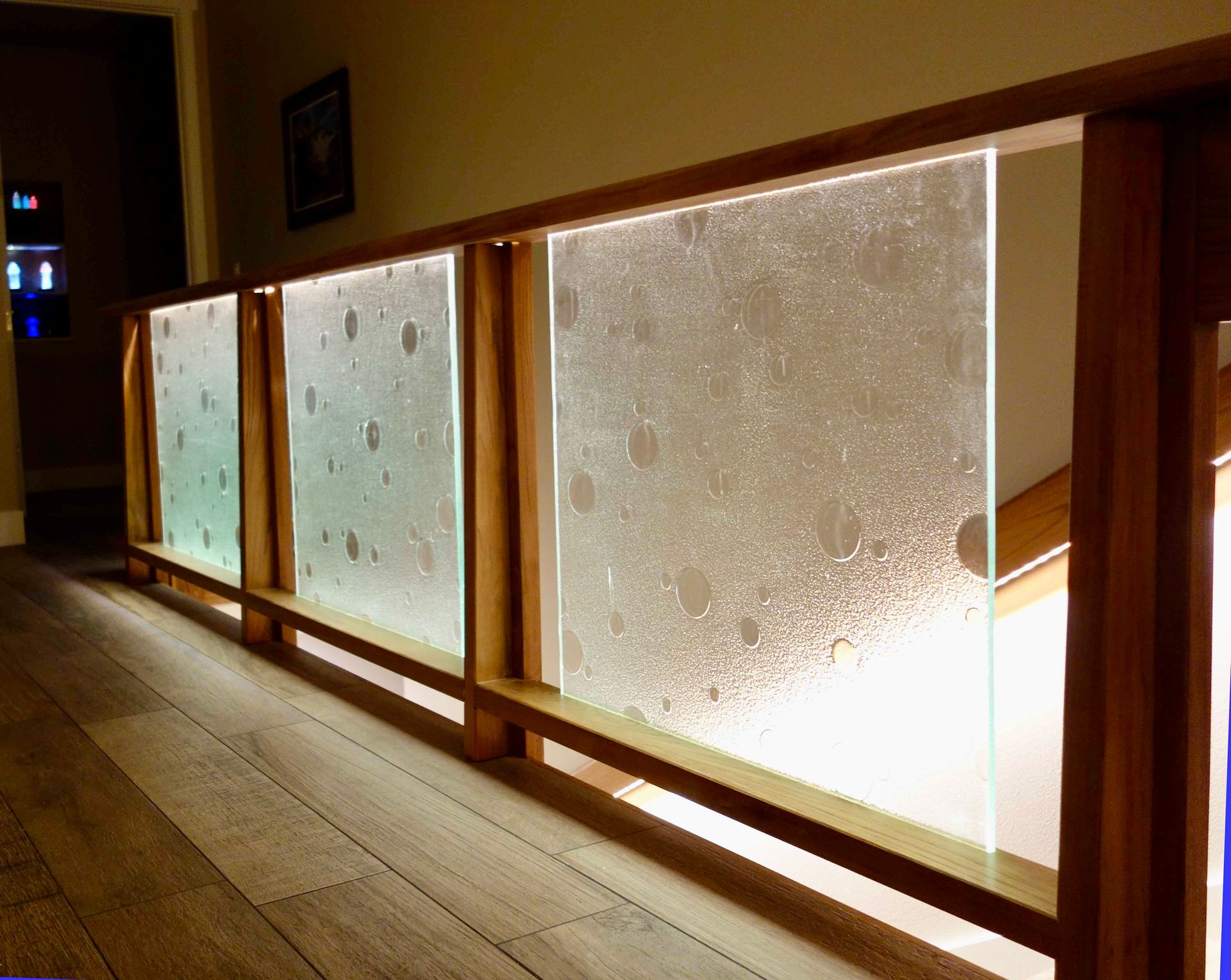 A wooden stair railing with illuminated, textured glass panels featuring a subtle, circular pattern.