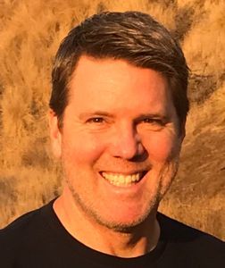 Smiling man with short gray hair, wearing a black shirt, in front of yellow-brown tall grass.