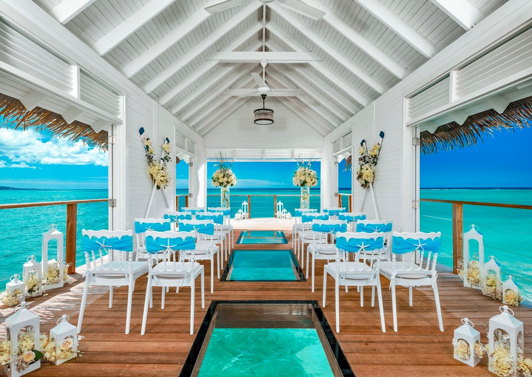 An overwater wedding pavilion with rows of white chairs, blue accents, and glass floor panels overlooking turquoise water.