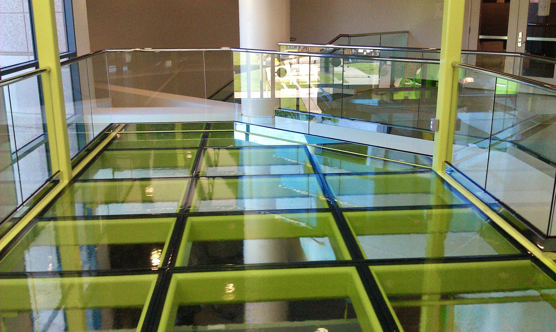 A glass floor walkway with a bright green metal frame, elevated above an open, modern interior space.