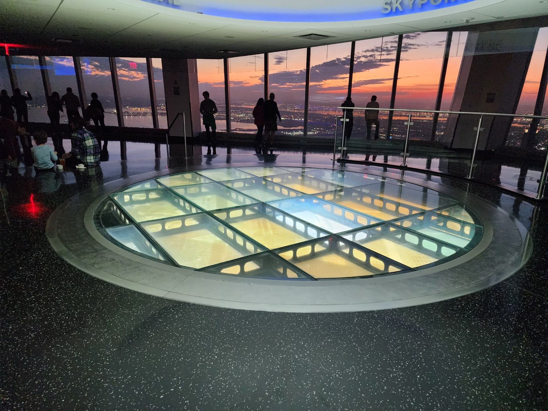 Glass floor observation deck with sunset view, people silhouetted against the sky.