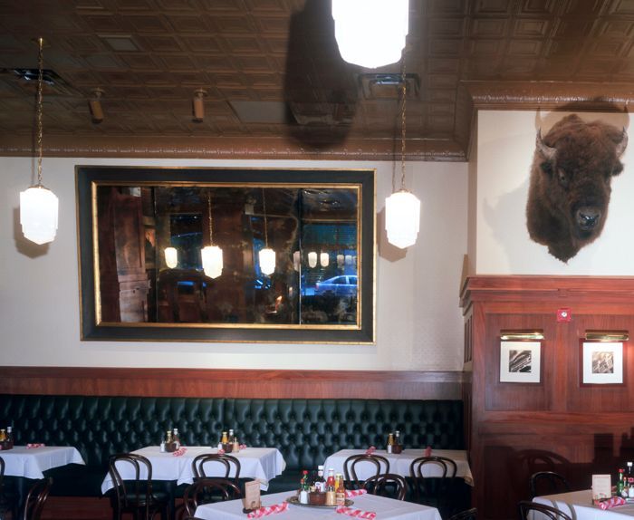 Restaurant interior with tables, tufted green booth, antique mirror, bison head, and hanging lights.