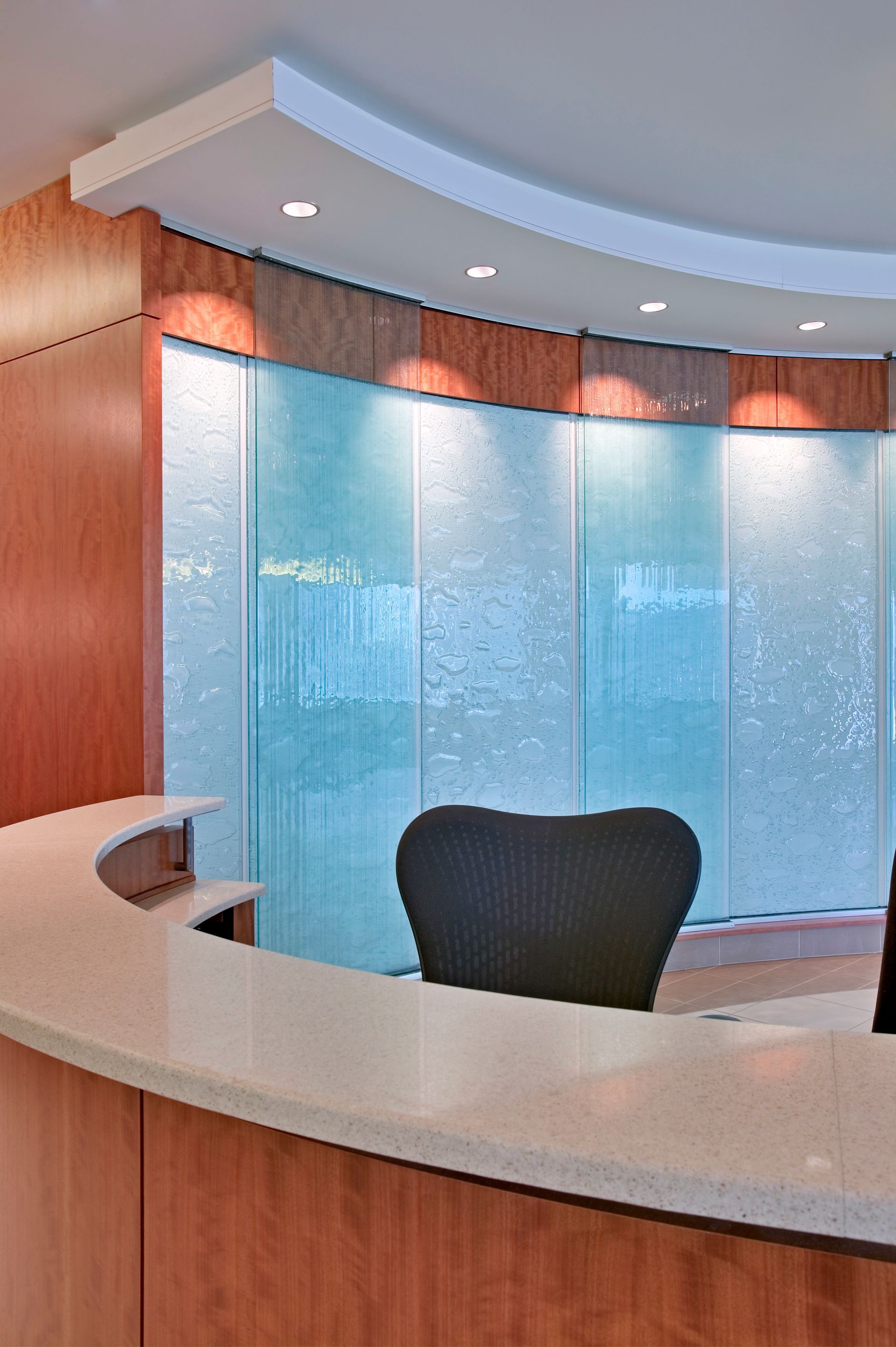 Modern lobby reception desk with curved wood counter, blue glass wall, and ceiling lights