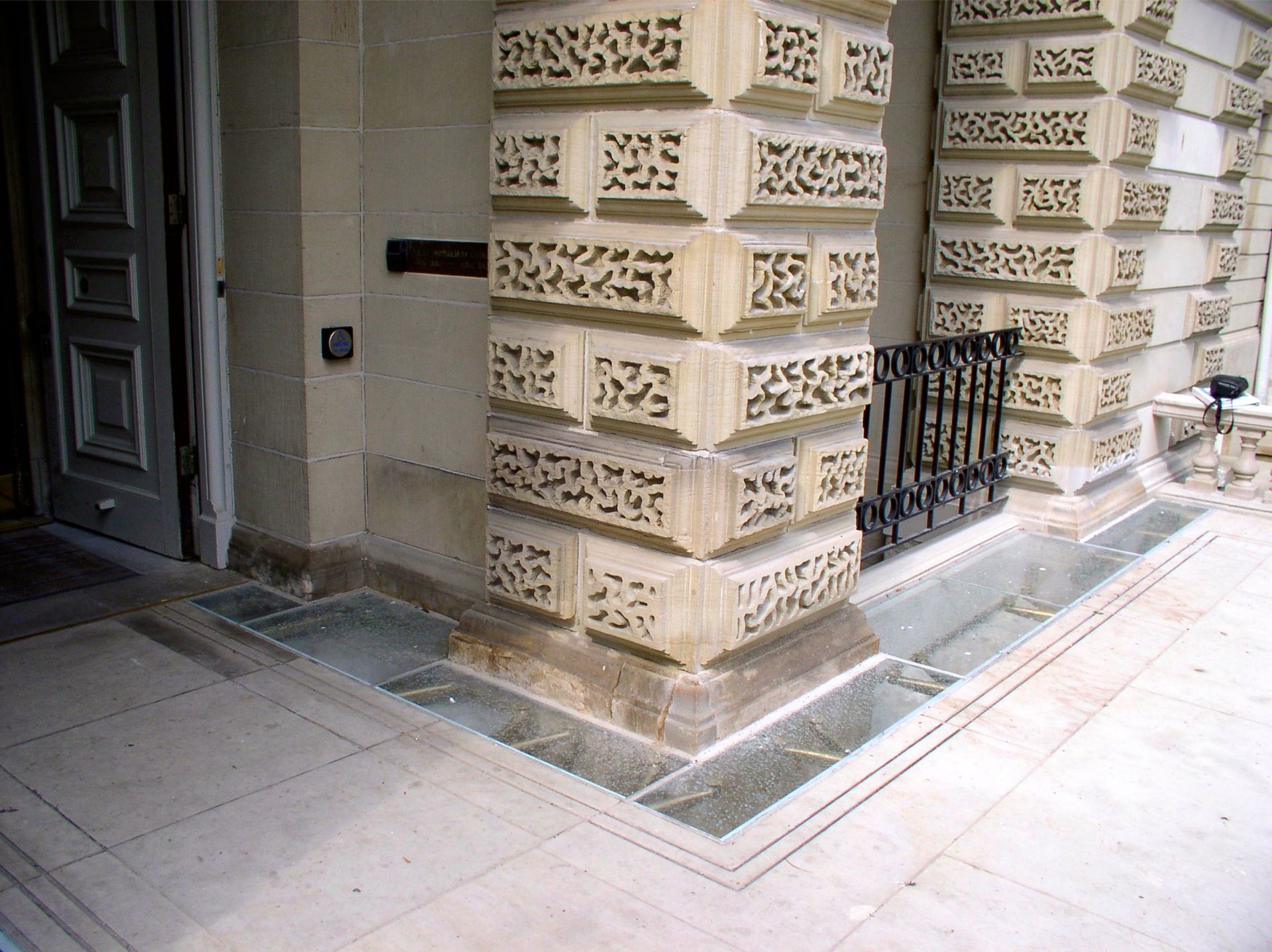 Beige stone building entrance with patterned pillar and a small window with a black metal fence.