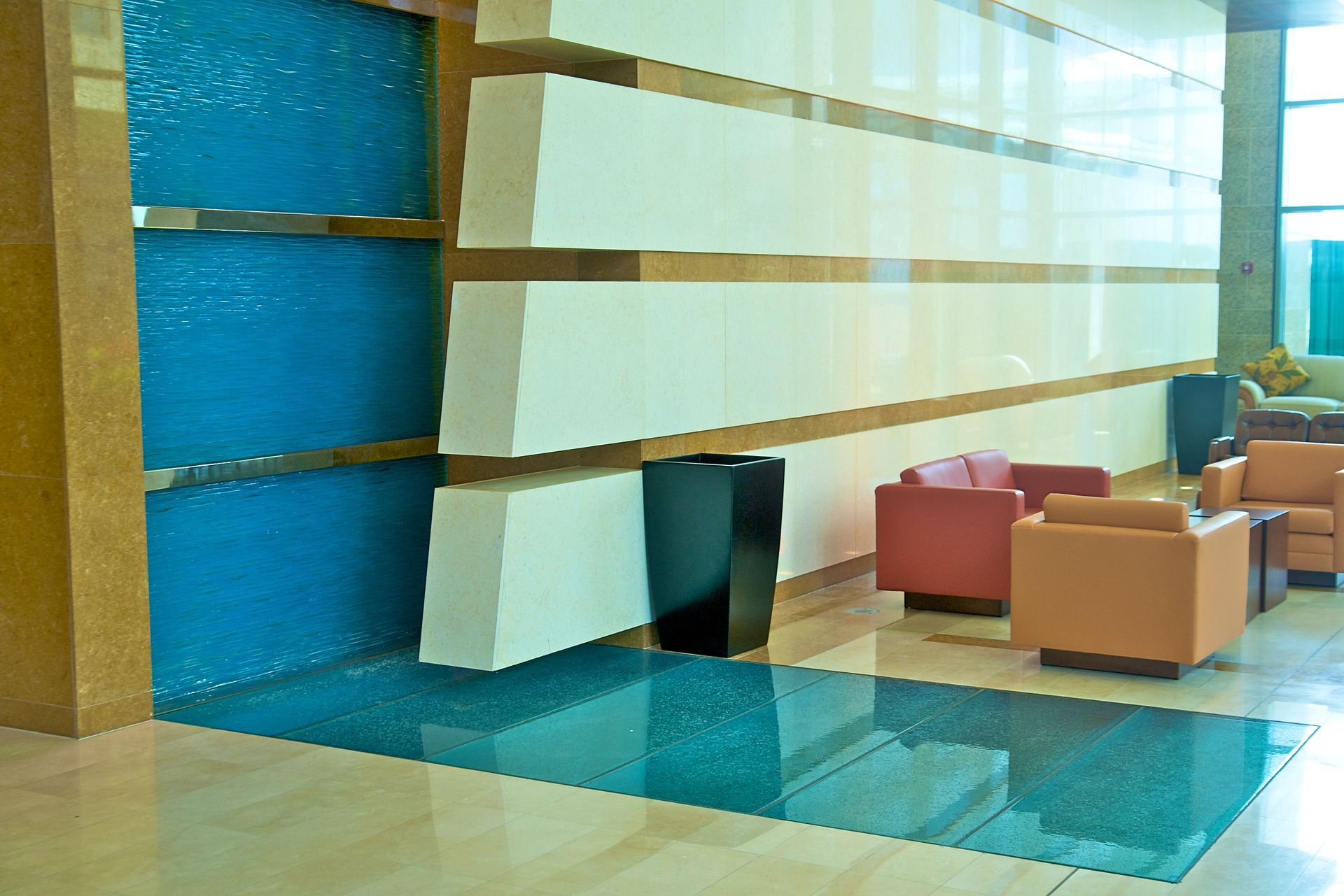 Blue water wall feature in a lobby with tan and white walls, orange and red seating, and a reflective floor.