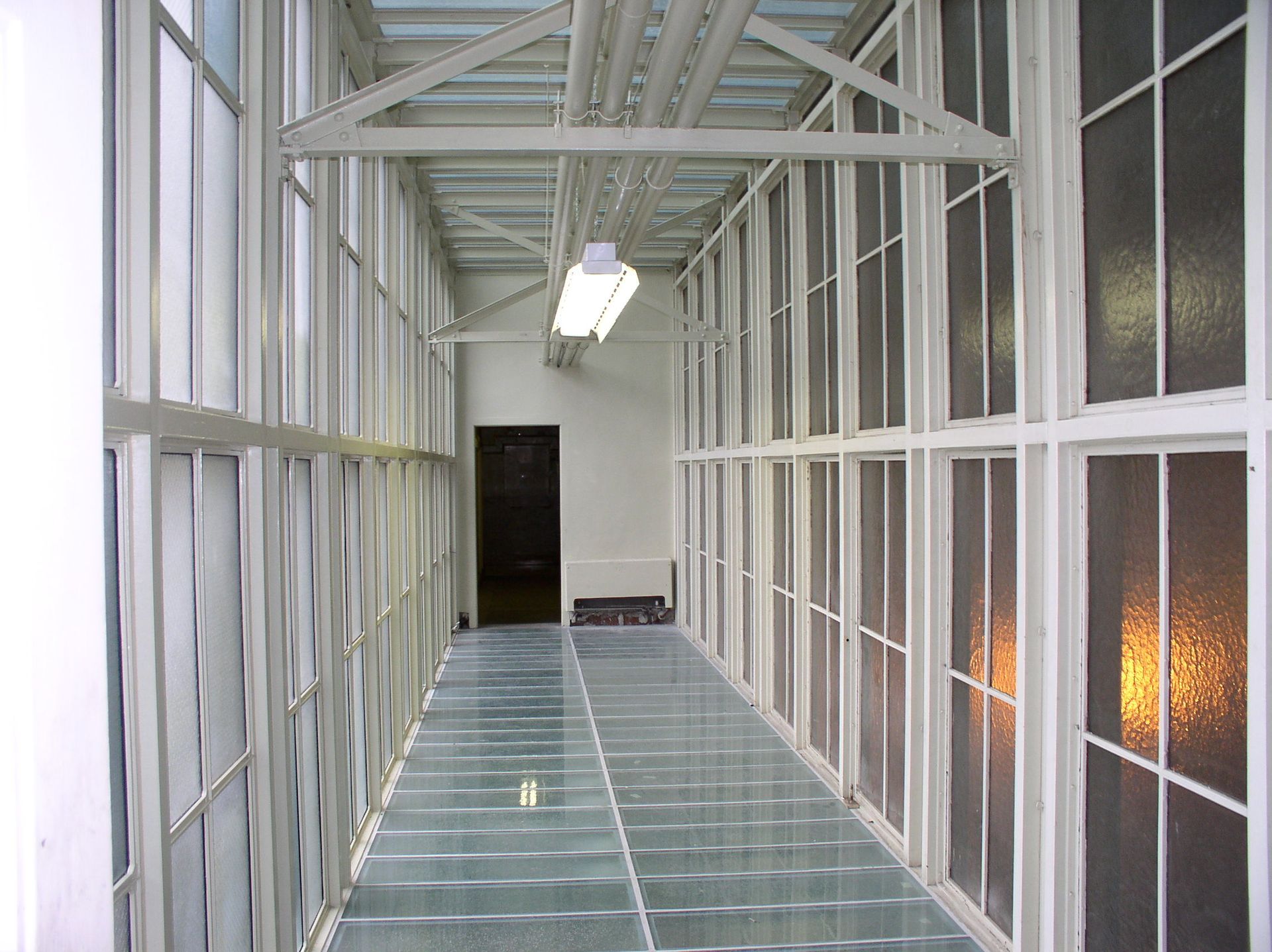 Glass walkway with white metal framing and translucent walls, leading to a doorway.