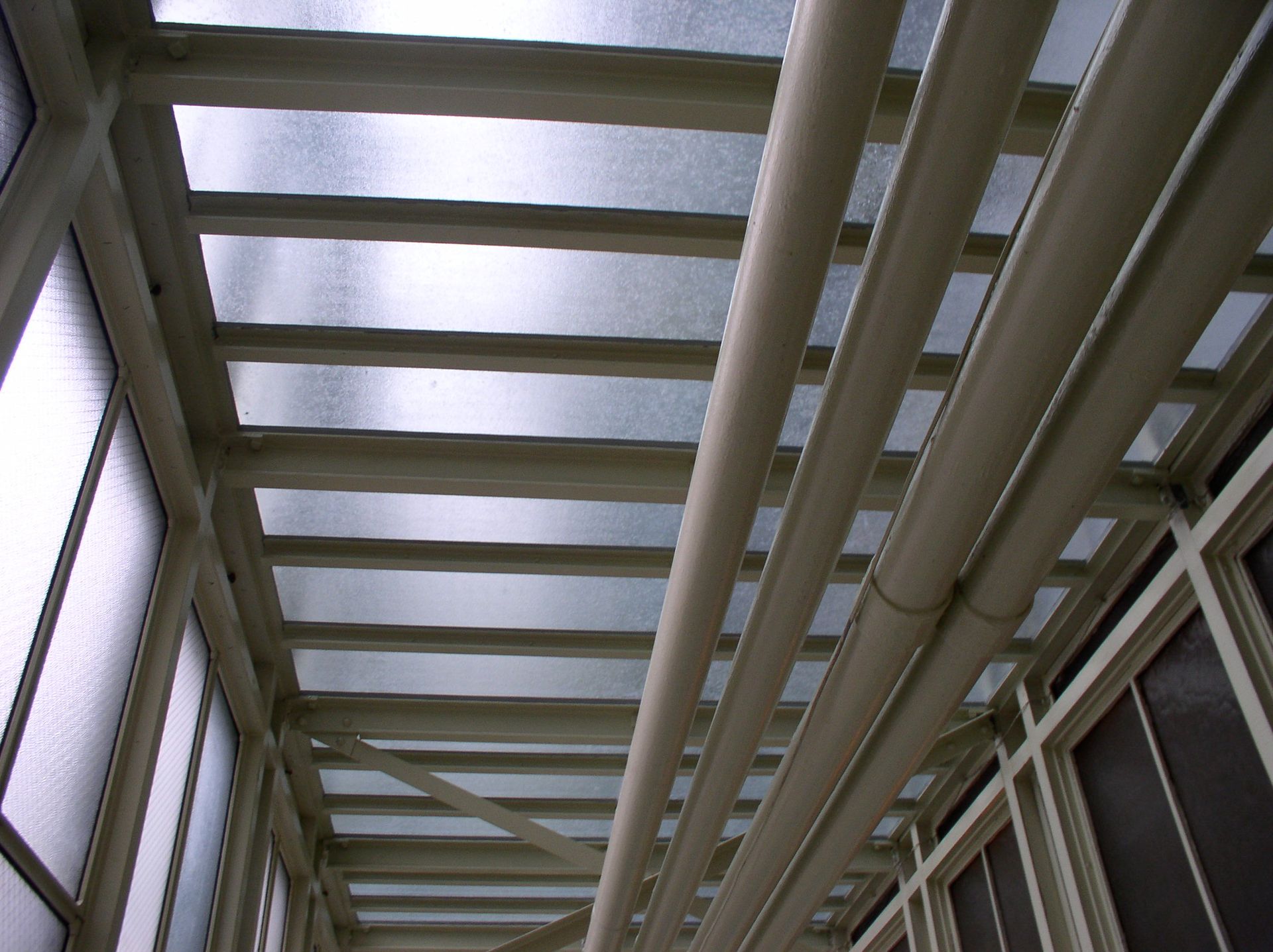 A glass ceiling with white beams and pipes, part of a walkway.