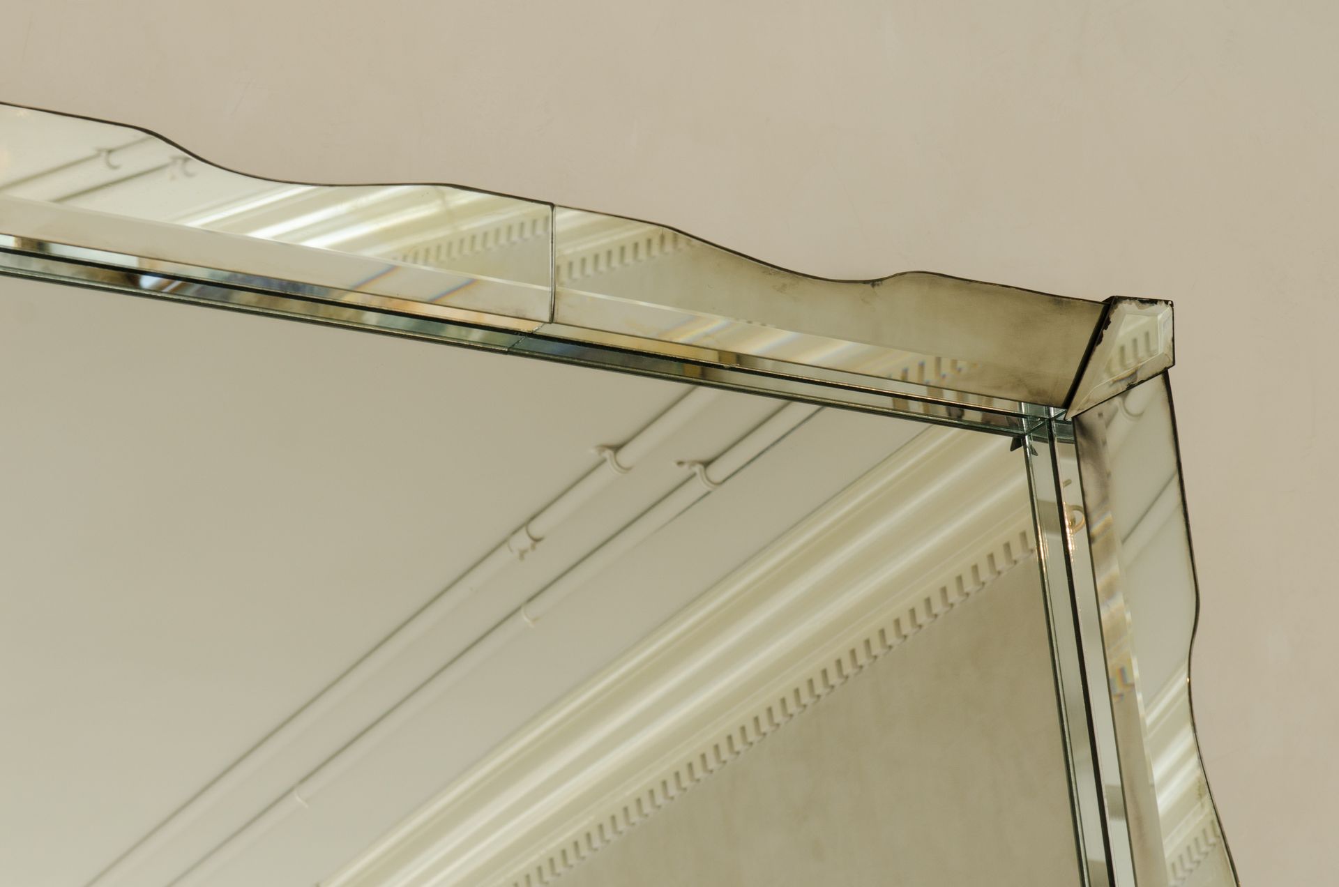 Close-up view of a glass-framed mirror with a decorative, wavy top edge, mounted on a cream-colored wall.
