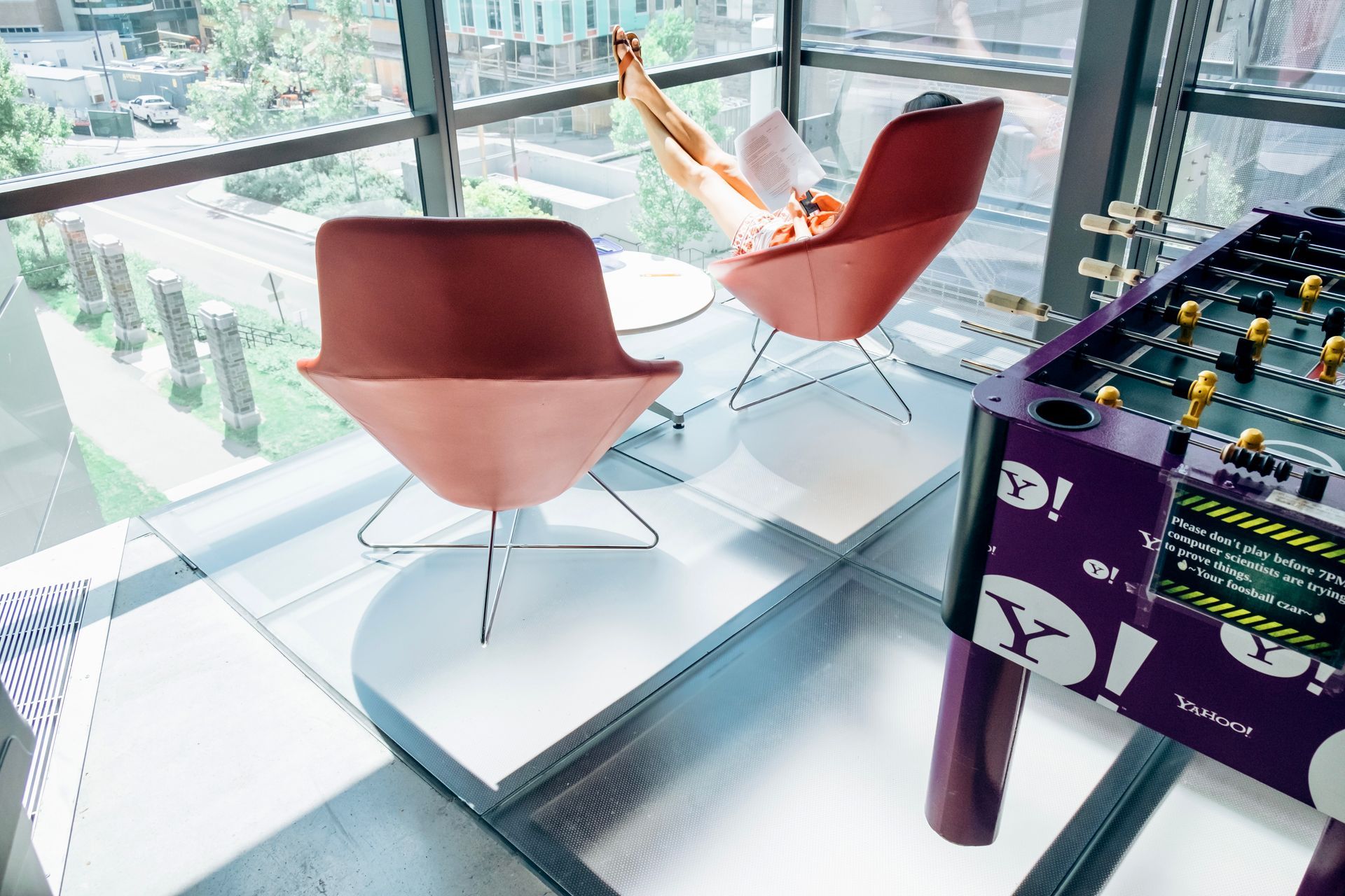 A person relaxes in a red chair by a window, near a foosball table in a brightly lit room.