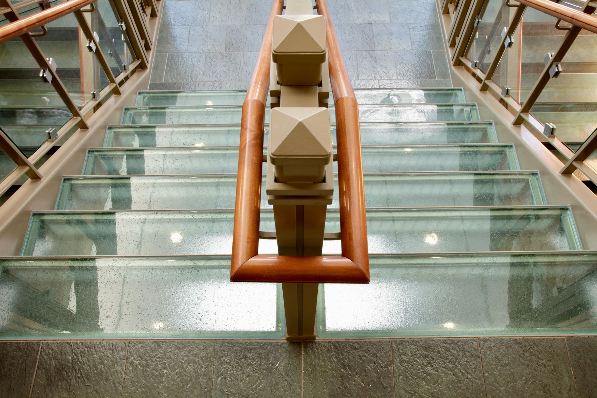 A top-down view of a staircase with glass treads and wooden railings, descending toward a stone floor.