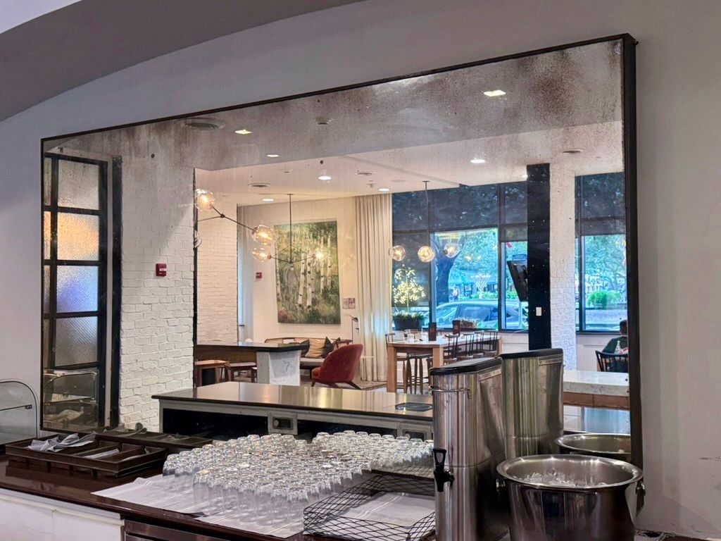 A large, vintage-style wall mirror reflects a bright restaurant interior with tables, a painting, and light fixtures.