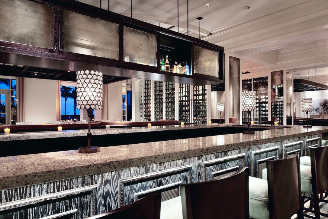 Bar with a long counter, brown chairs, and a glass-fronted shelf holding liquor bottles; interior setting.