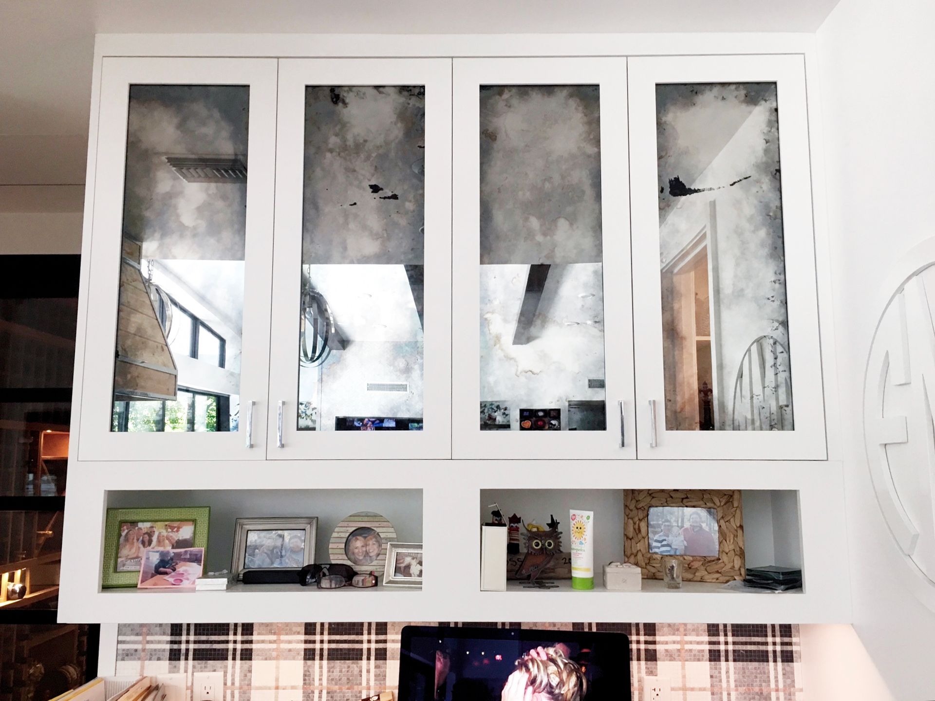 White cabinets with antique mirrored doors above a shelf displaying photos and skincare, over a television.