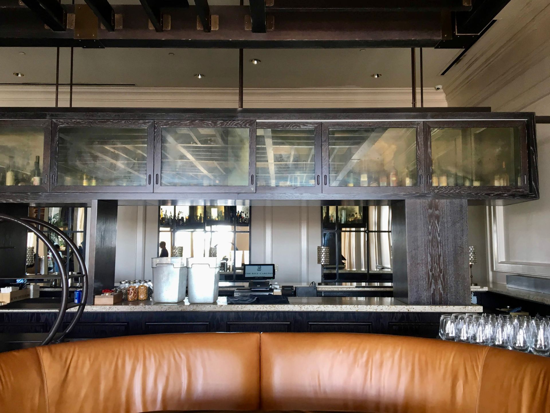 Bar interior with brown leather booth, dark wooden bar, and antique mirror glass-fronted cabinets displaying bottles.