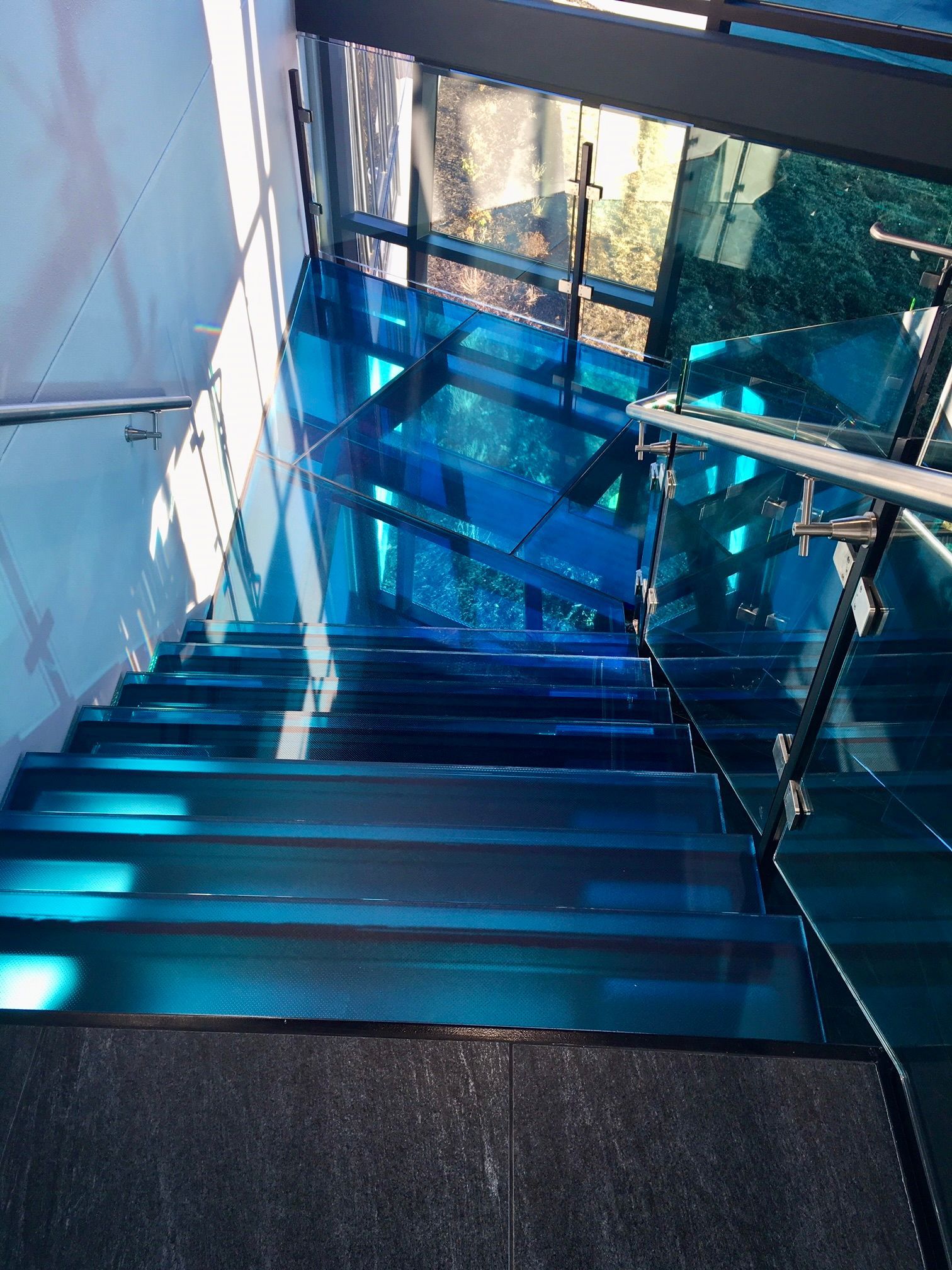 Blue glass staircase with metal railing; sunlight casts shadows.