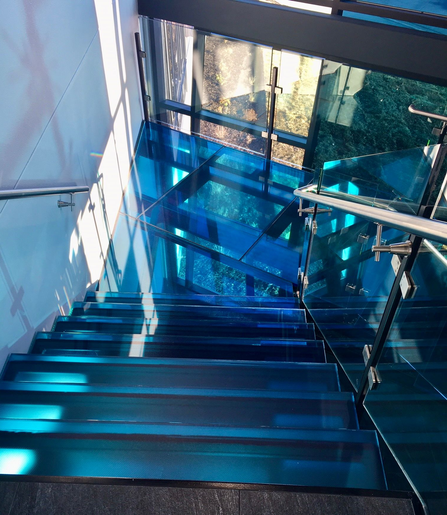 Blue glass treads staircase with chrome railing, sunlight creating shadows.