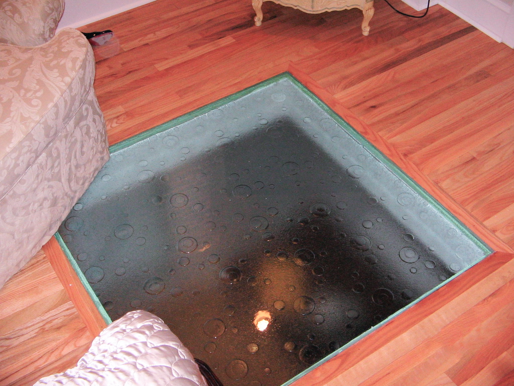 A glass floor panel inset into a wooden floor, revealing a dimly lit, dark space below.