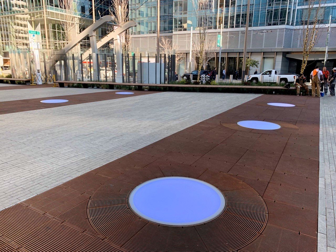 Exterior shot of a plaza with brown and grey paving stones and circular glass floor accents.