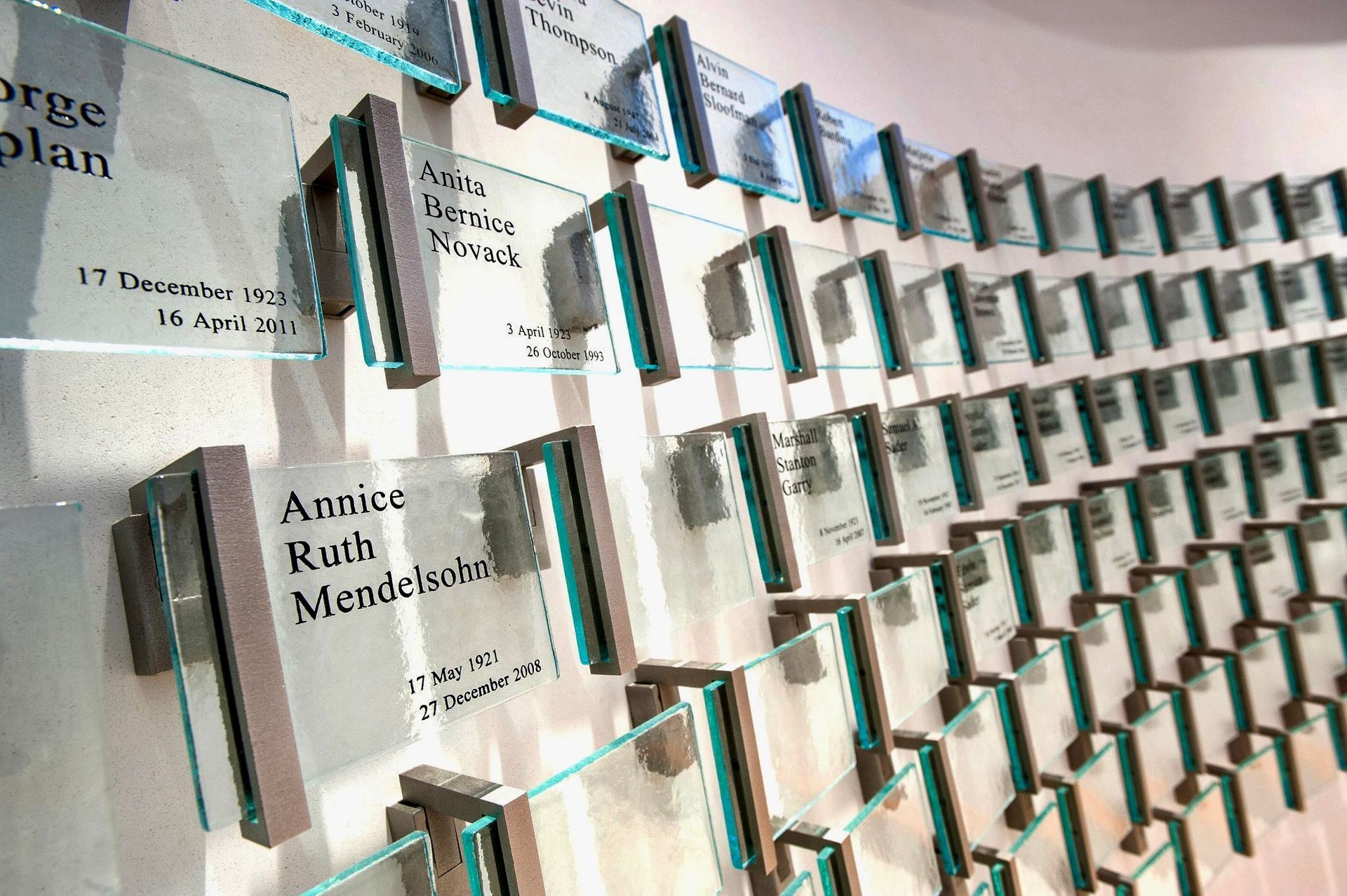 A curved wall displaying rows of individual glass memorial plaques with names and dates, mounted on metal brackets.