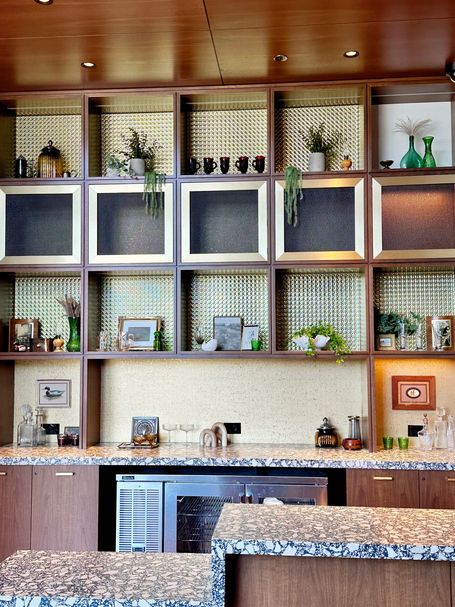Wooden bar with shelves containing decor and drinks, granite countertop, mini-fridge.