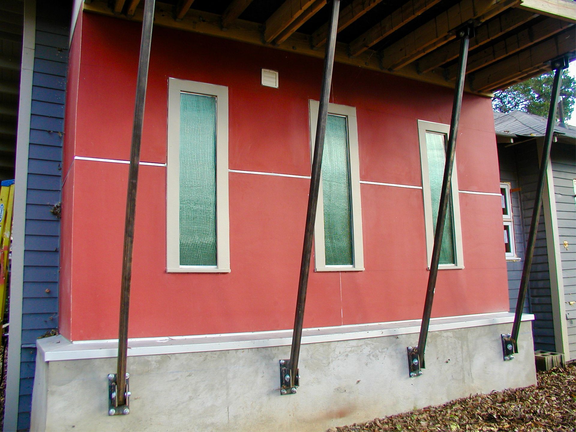 A red exterior wall with three vertical windows, braced by diagonal metal supports anchored to a concrete foundation.