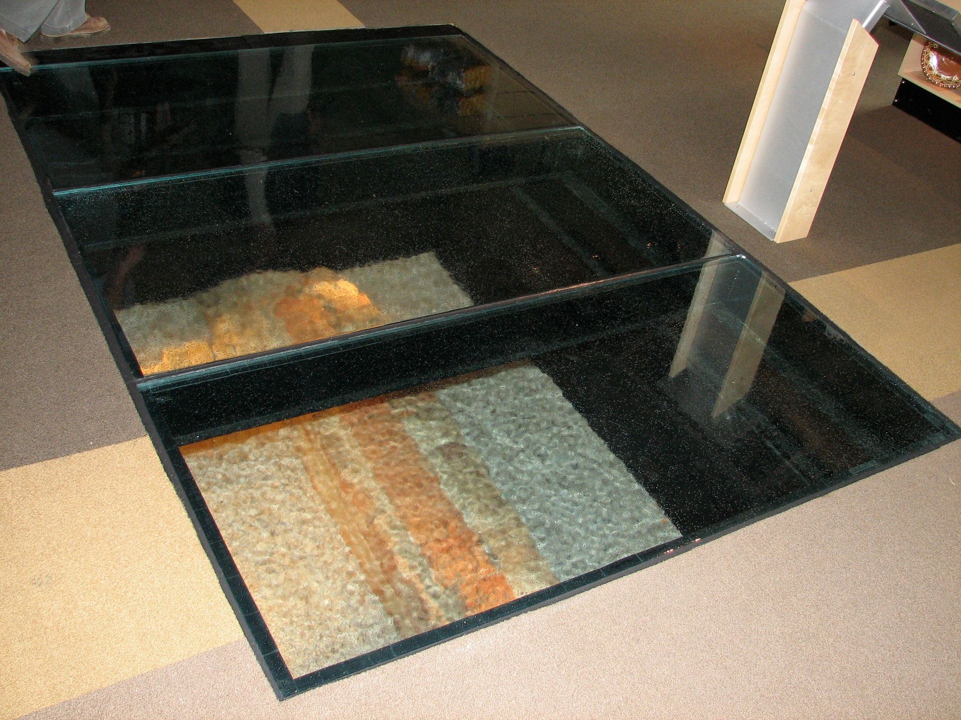 A glass panel floor embedded in a carpeted room, revealing archaeological remains underneath.