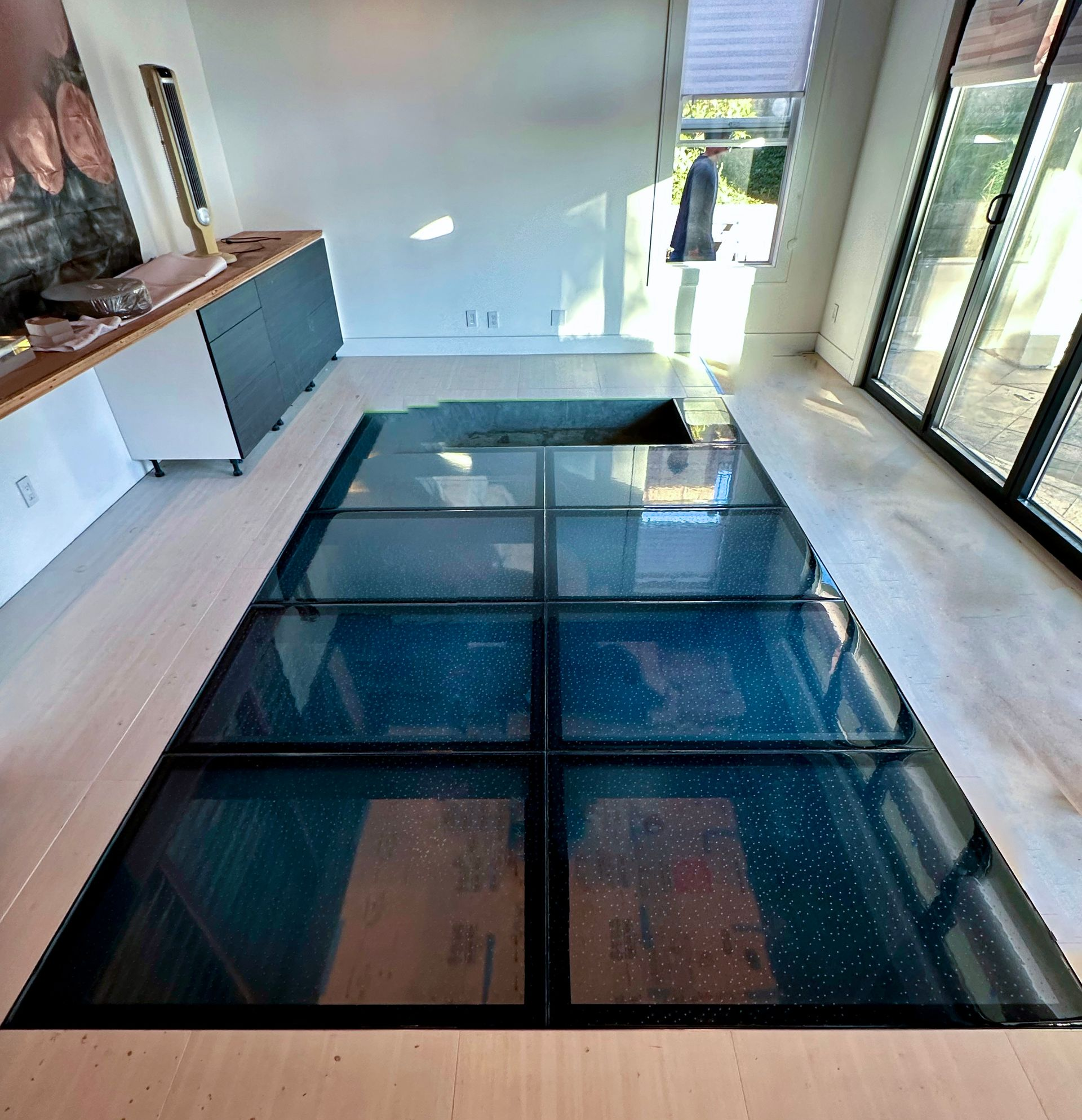 Glass floor panels in a modern room with a dark frame. Sunlight streams through.