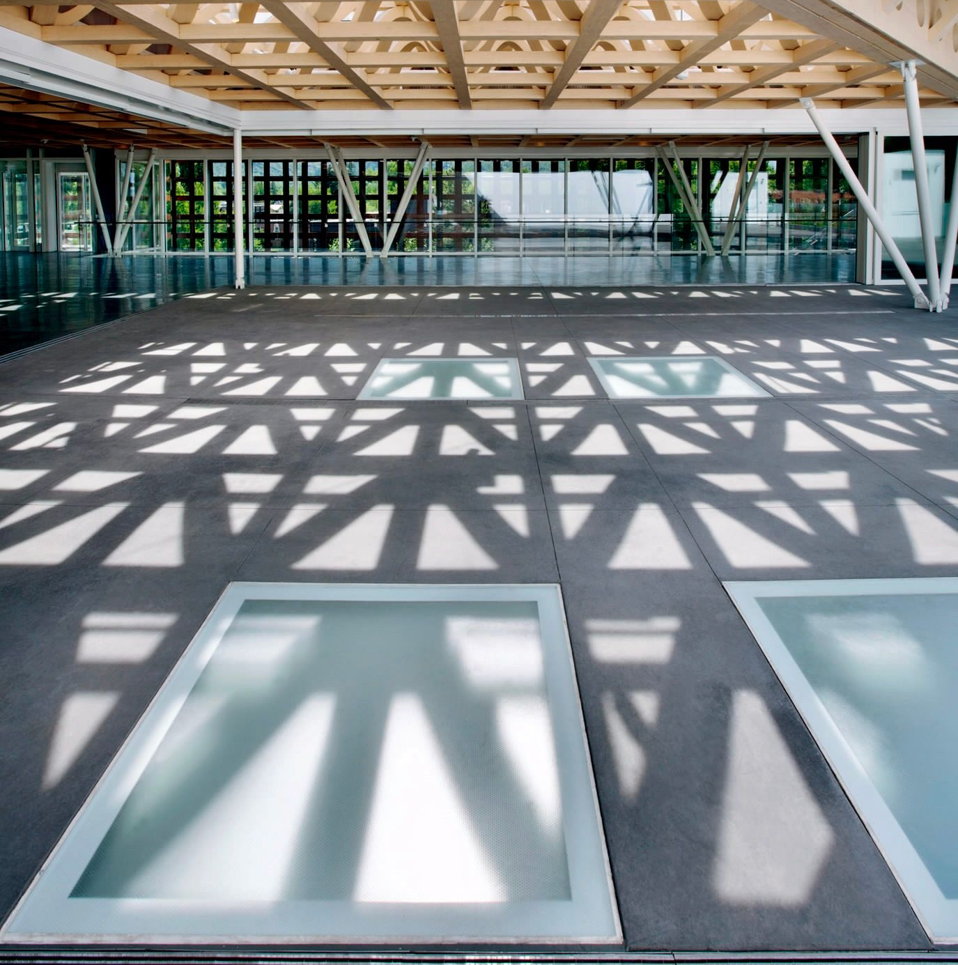 Interior view of a modern building with a geometric wooden ceiling casting intricate shadows on a floor with glass panels.