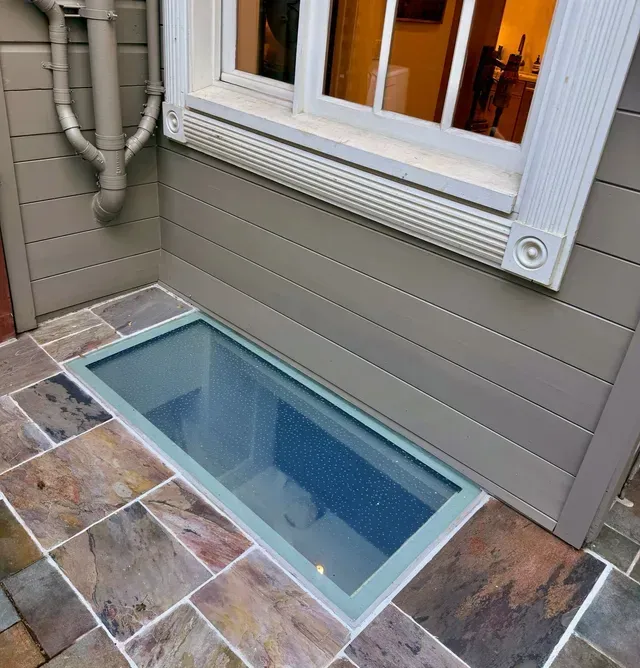Window well with glass floor panel inset in stone patio, below a window on a gray building.