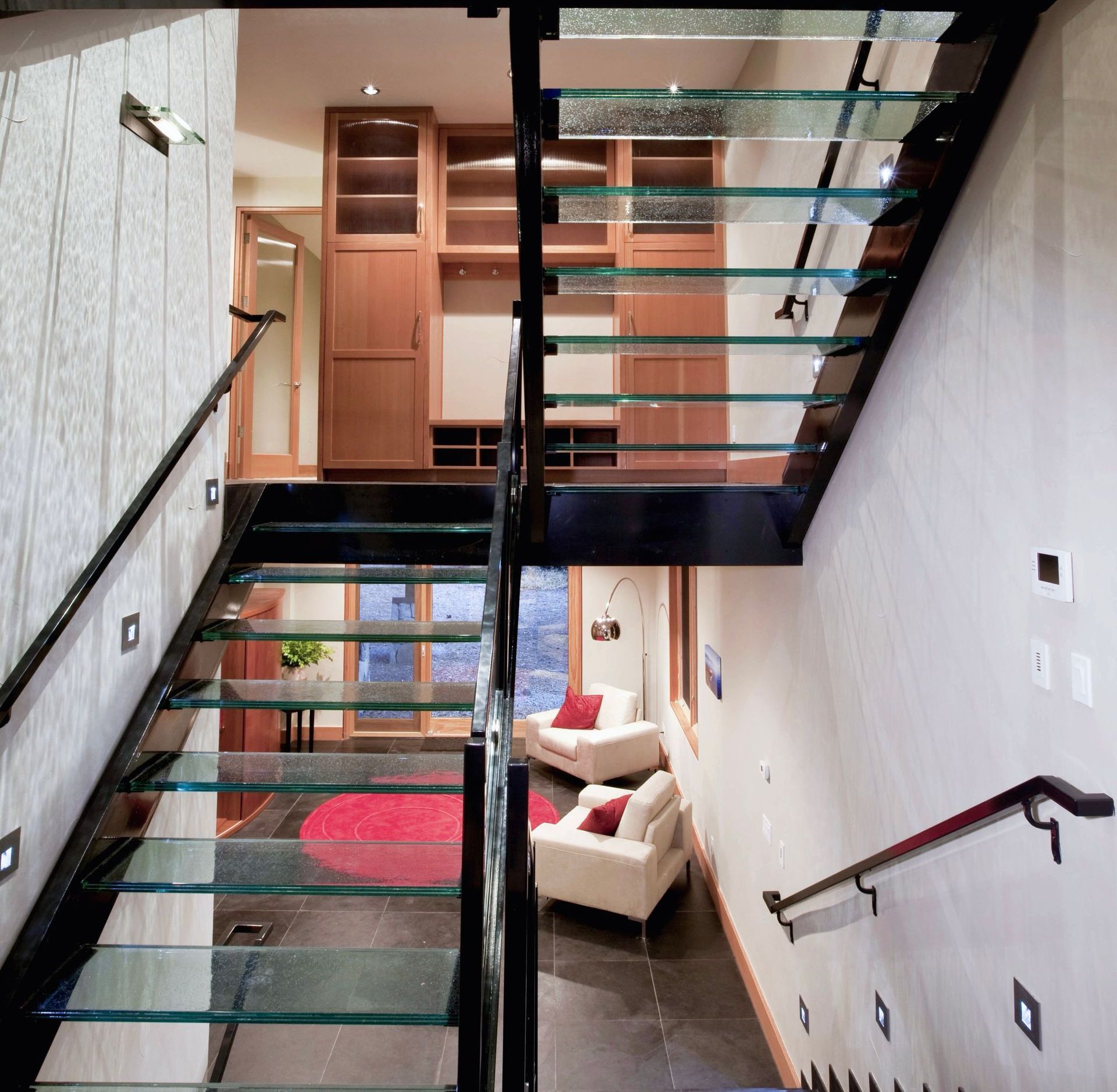 Modern staircase with glass steps and black metal frame, leading to a seating area with white chairs and a red rug.
