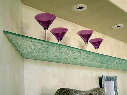 Four purple martini glasses displayed on a thick, textured, pale green glass shelf mounted on a beige wall.