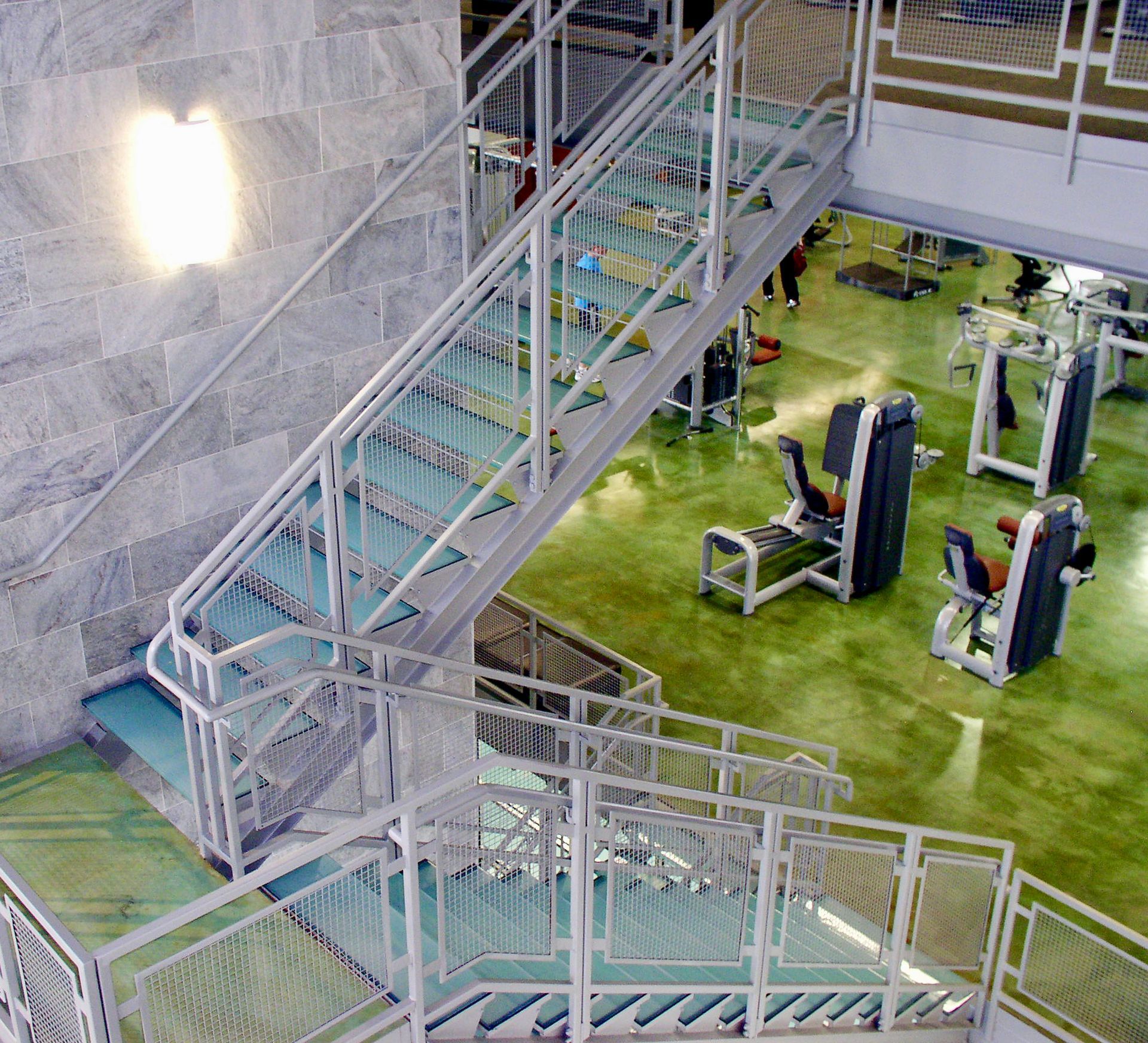 Glass staircase overlooking a gym with exercise equipment. White railings, teal-colored flooring, and green gym floor.