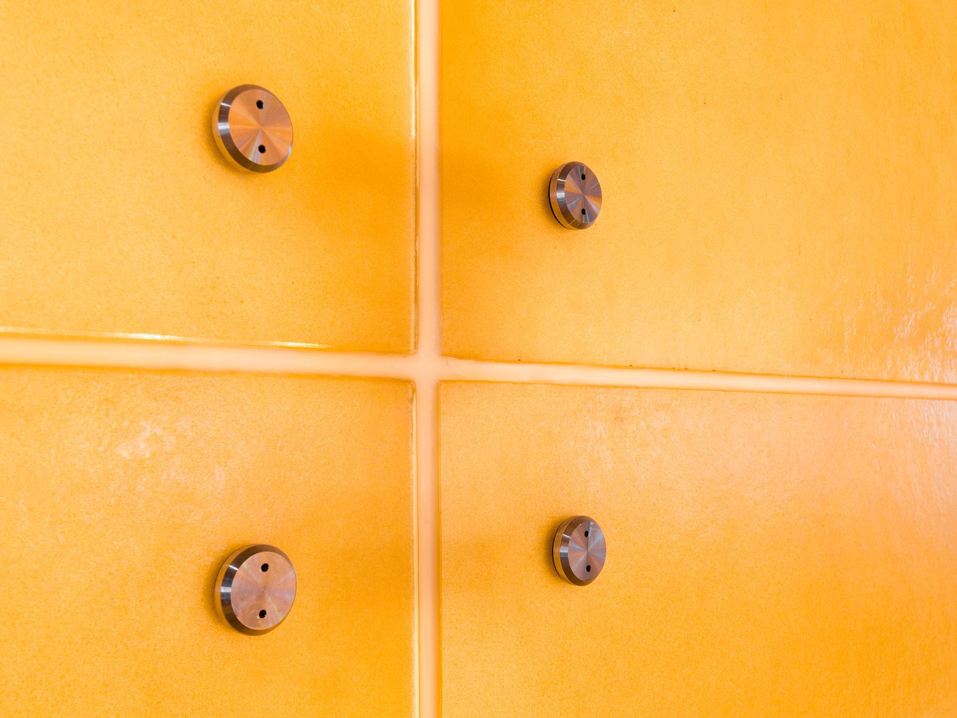 Four metallic circular fasteners mounted on bright yellow, tiled wall panels meeting at a cross-shaped grout line.