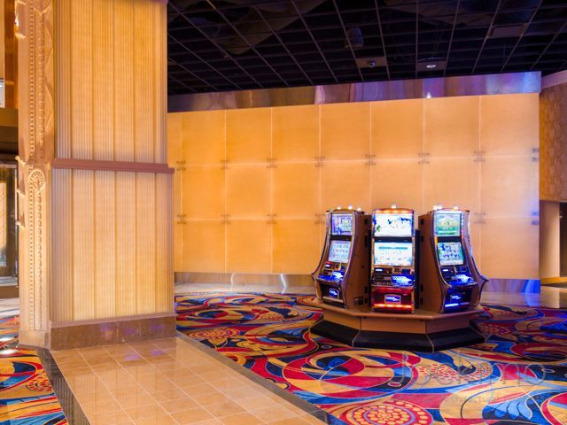 A cluster of three slot machines sits on a patterned carpet in a brightly lit casino lobby with paneled walls.