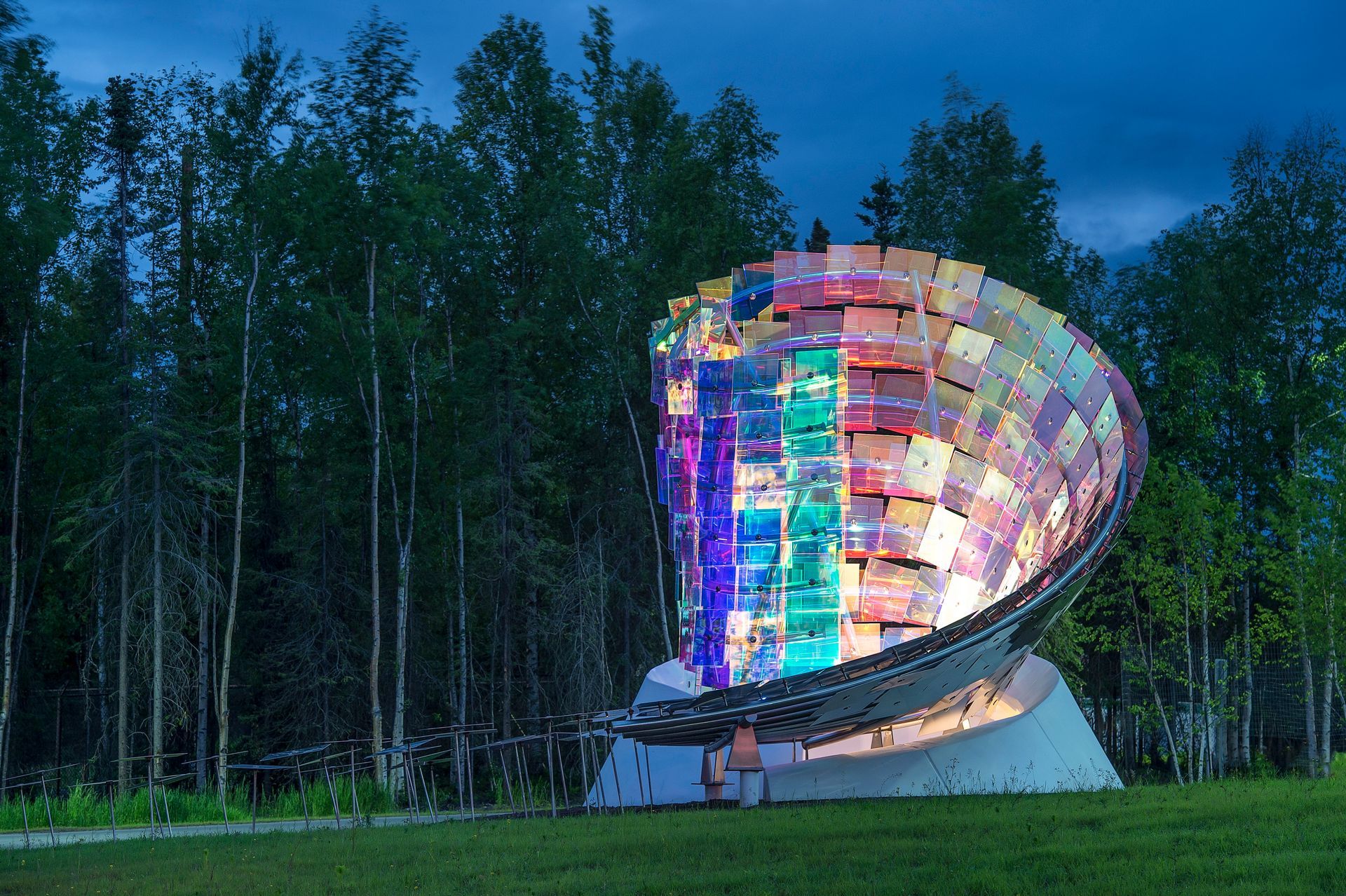 Colorful illuminated satellite dish sculpture in a grassy field at dusk