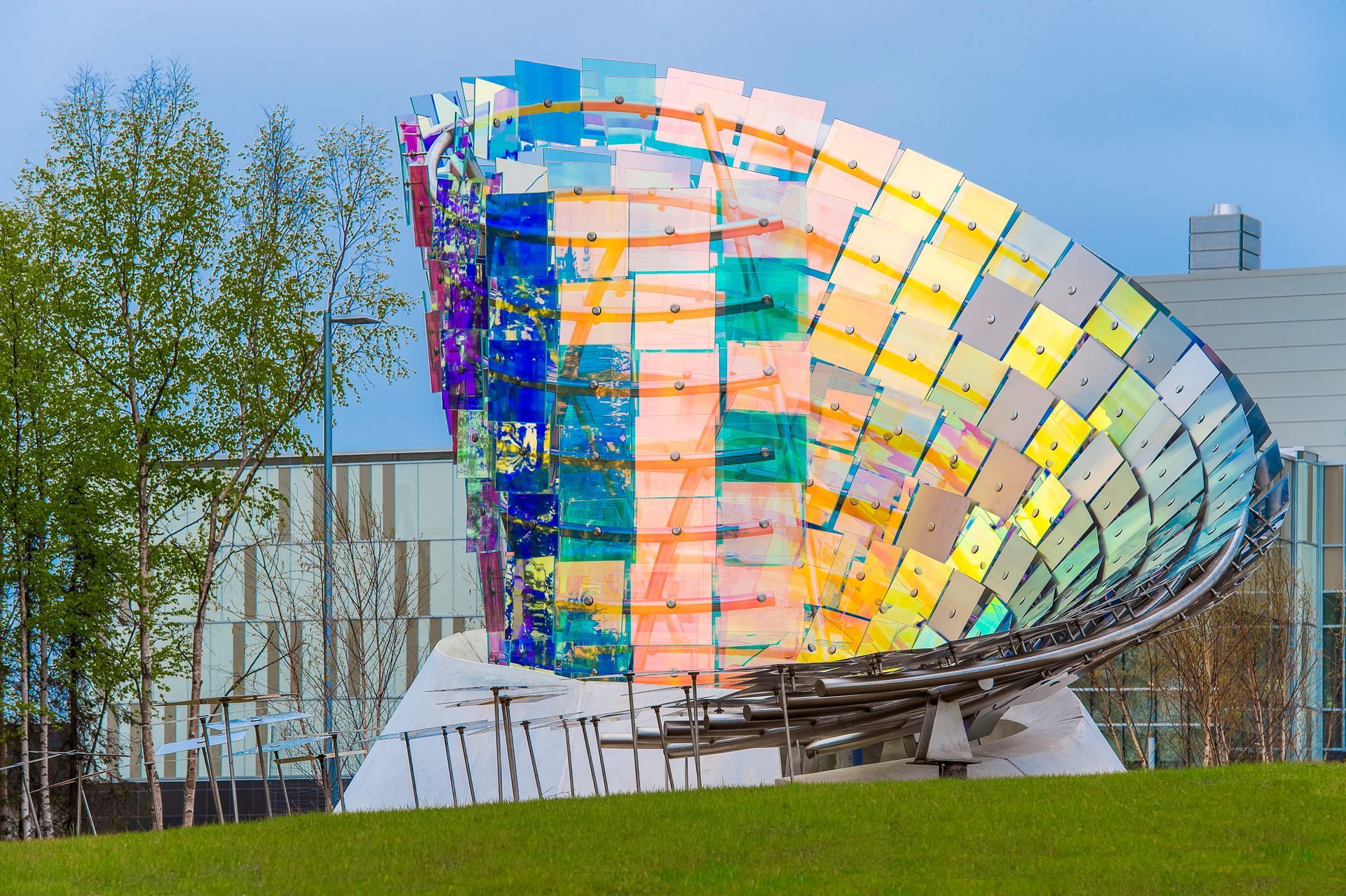 Colorful curved glass art sculpture in a grassy outdoor plaza with trees and a building in the background