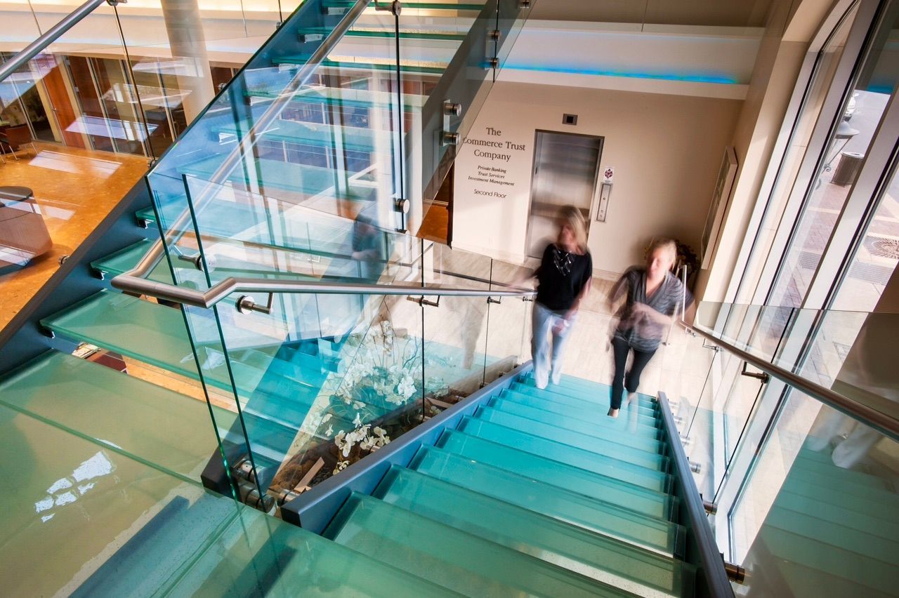 Two women descending glass stairs; bright interior, sunlight.