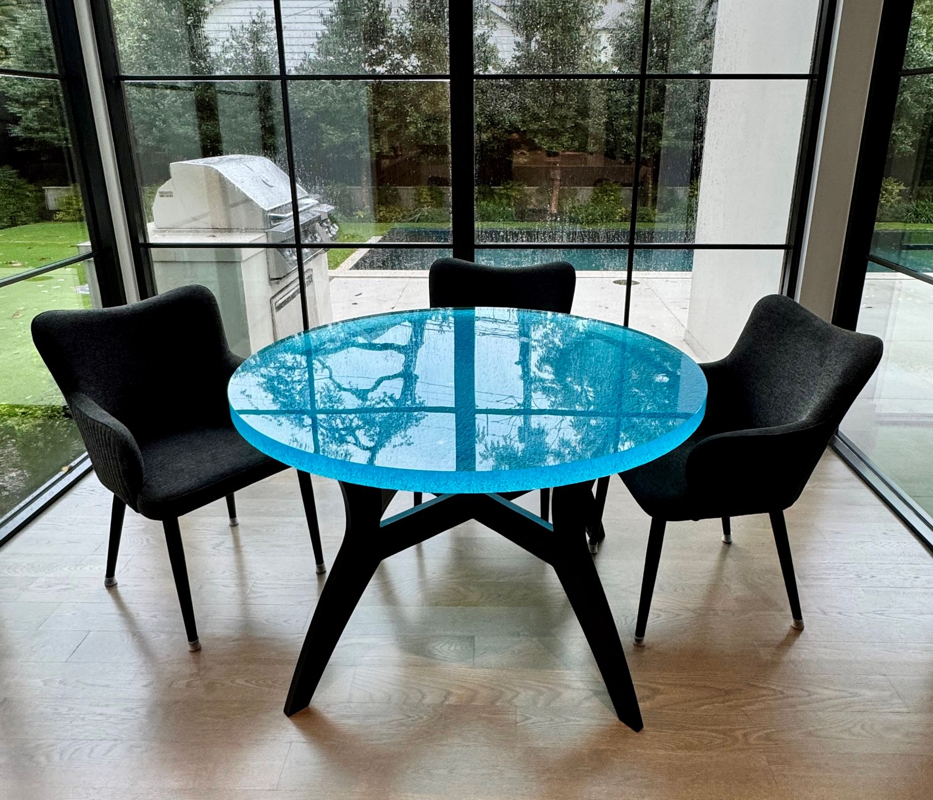 A round, turquoise-colored glass table with three black chairs, set near large windows overlooking a backyard.