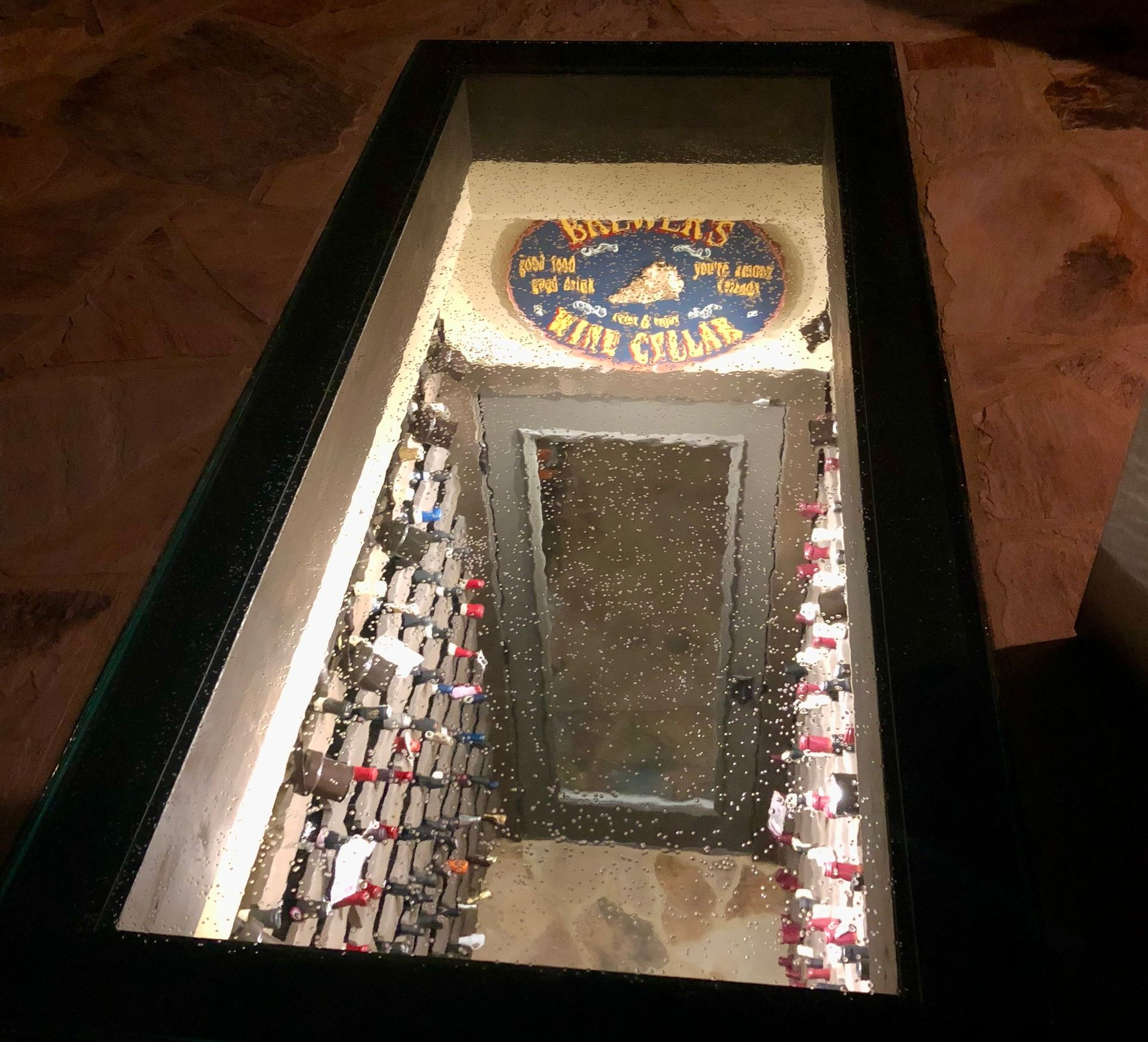 A view looking down through a glass floor panel into a lit wine cellar containing rows of wine bottles on shelves.