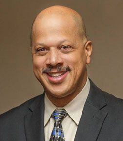Bald man in a suit smiles; tan skin, brown mustache, patterned tie, neutral background.