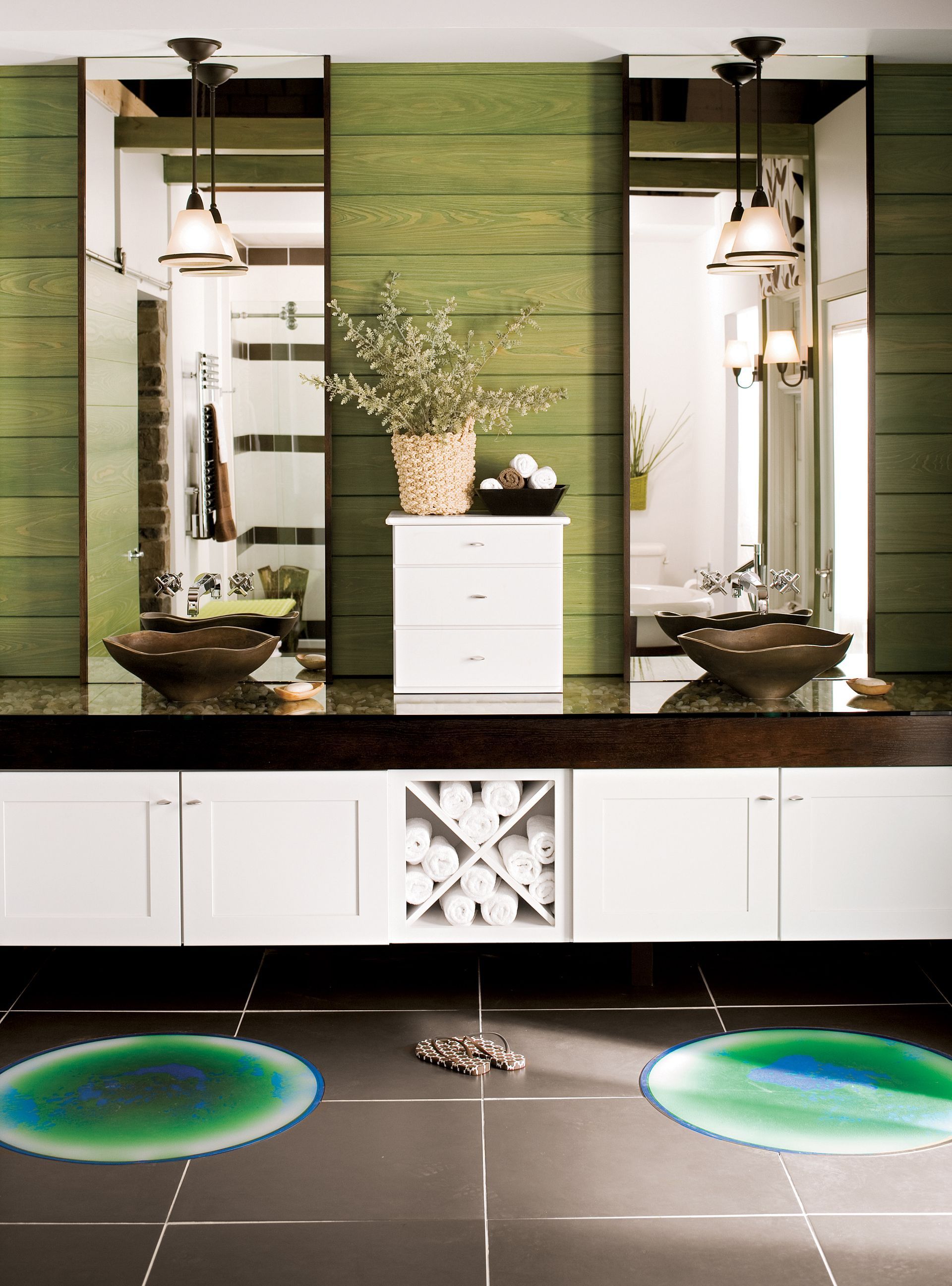 Modern bathroom with white cabinets, dark countertops, twin sinks, green wood-paneled walls, and two colorful circular rugs.