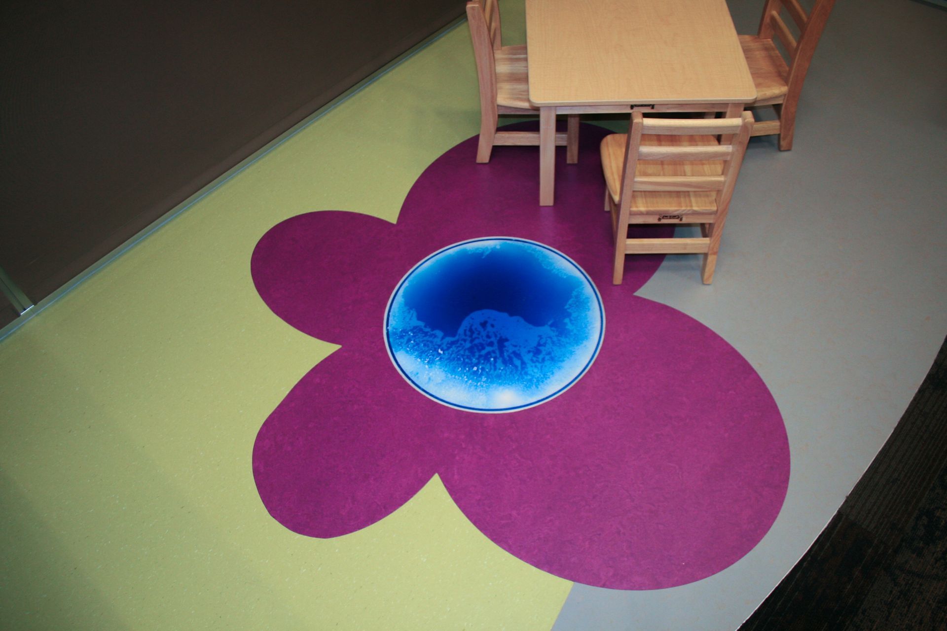 A purple flower-shaped floor mat with a blue, water-like center, placed beside a wooden table and chairs in a room.