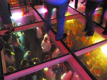 People dancing on a glass dance floor above a crowd in a brightly lit club.