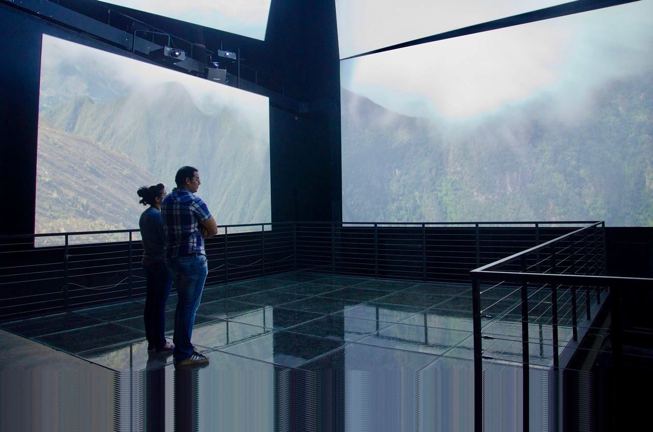 Two people stand on a reflective glass platform inside a dark room, viewing large screens showing misty mountain scenery.